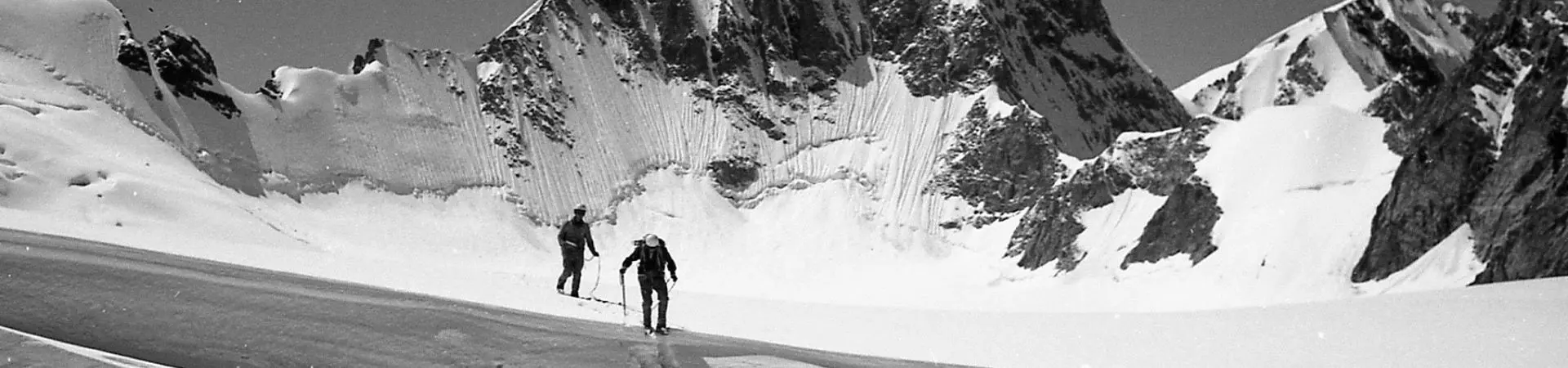 Vier bergbeklimmers in zware winteruitrusting staan op de besneeuwde top van een berg, naast een stapel ski’s en uitrusting, onder een helderblauwe hemel.