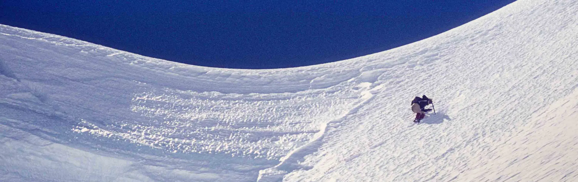Twee bergbeklimmers in zware winteruitrusting, met een skibril en handschoenen, staan even stil op een besneeuwde berghelling. De ene bergbeklimmer zit gehurkt en houdt een opgerold touw vast; de ande