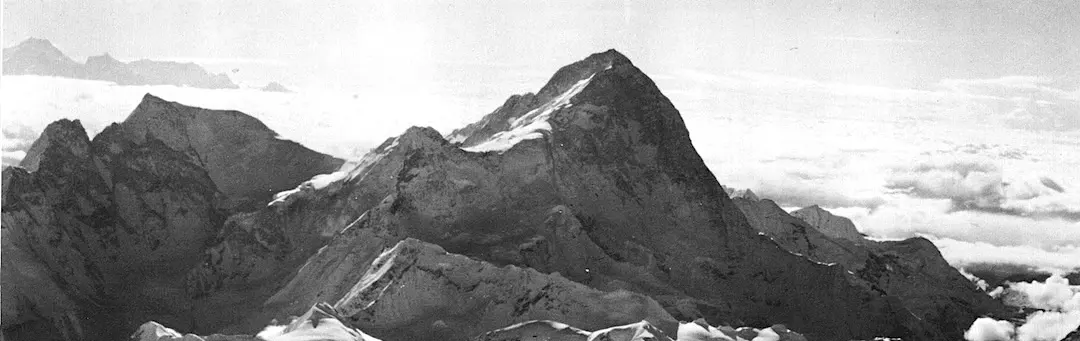 Een bergbeklimmer staat op een besneeuwde bergkam en kijkt uit over een indrukwekkende bergketen met steile, met sneeuw bedekte toppen die in de verte scherp omhoog rijzen onder een bewolkte hemel.