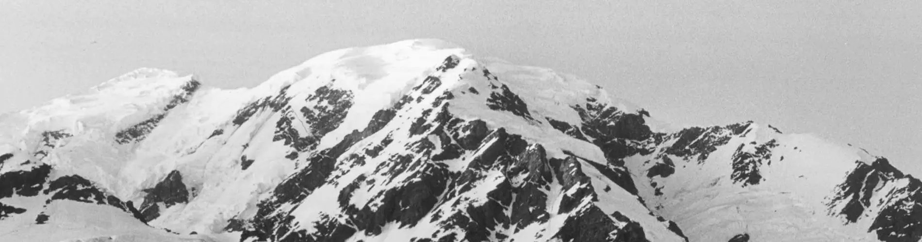 Op een zwart-witfoto is een met sneeuw bedekte berg te zien met grillige, rotsachtige uitlopers en diepe spleten. Sneeuw en ijs bedekken de hellingen en vormen een contrast met de donkerdere rotsen er