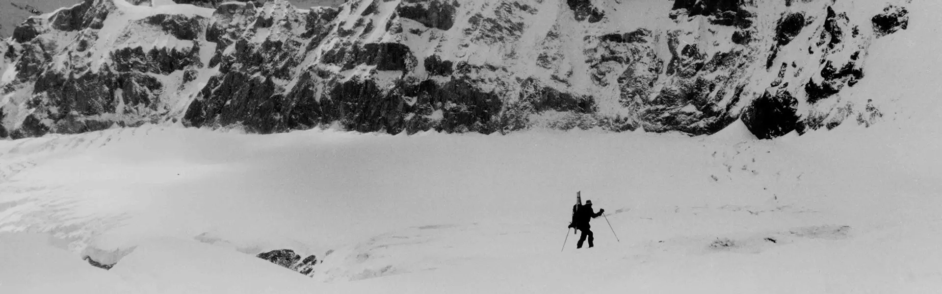 Drie bergbeklimmers, gekleed in dikke winterjassen, handschoenen, gezichtsmaskers en skibrillen, zitten dicht bij elkaar in de sneeuw en houden een vlag vast. Hun gezichten worden gedeeltelijk aan het