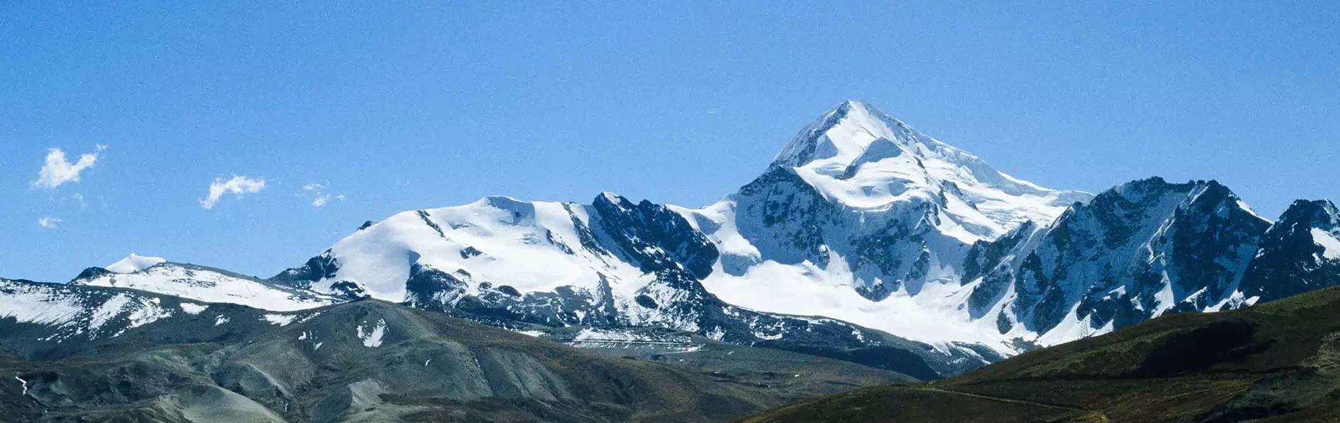 Twee bergbeklimmers zitten uit te rusten op een besneeuwde berghelling onder een strakblauwe hemel; ze dragen een zonnebril, warme kleding en bergschoenen met beenkappen. Op de achtergrond strekken be