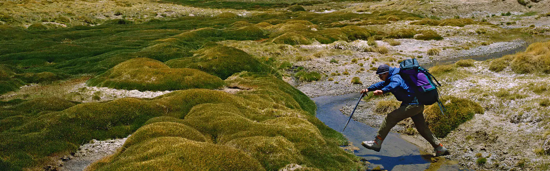 Een wandelaar met een rugzak gebruikt wandelstokken om een beekje over te steken in een met gras begroeide alpenvallei, omringd door ruige bergen en met sneeuw bedekte toppen onder een gedeeltelijk be