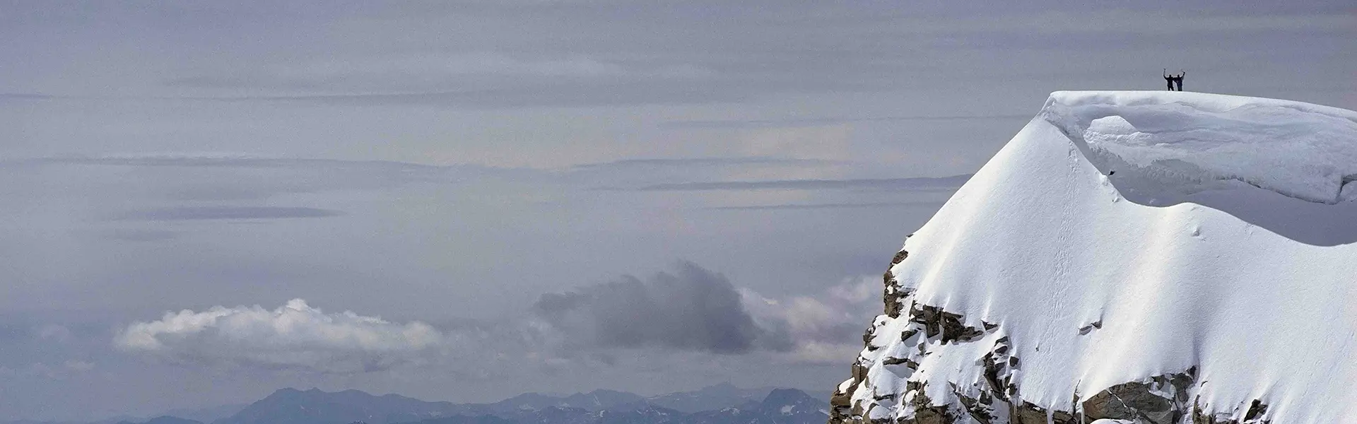Twee mensen staan op de besneeuwde top van een steile berg, omringd door ruige, met sneeuw bedekte bergen onder een bewolkte hemel.