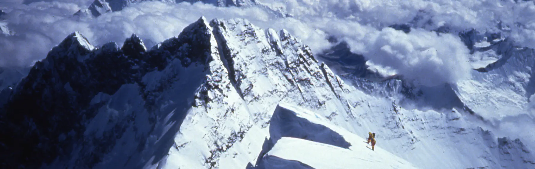 Drie bergbeklimmers in dikke rode en blauwe winterkleding en met skibrillen zitten samen op een besneeuwde berg; ze glimlachen en houden een klein Nepalees vlaggetje vast onder een strakblauwe hemel.