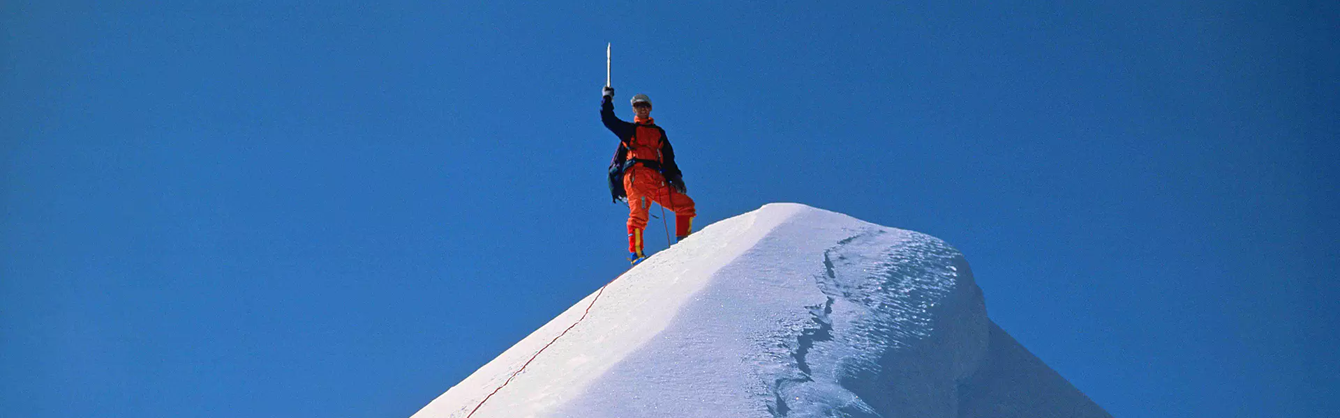 Een bergbeklimmer in oranje uitrusting staat triomfantelijk op een met sneeuw bedekte bergtop en houdt een ijsbijl omhoog, gericht naar de blauwe lucht. Achter hem loopt een rood touw langs de bergkam