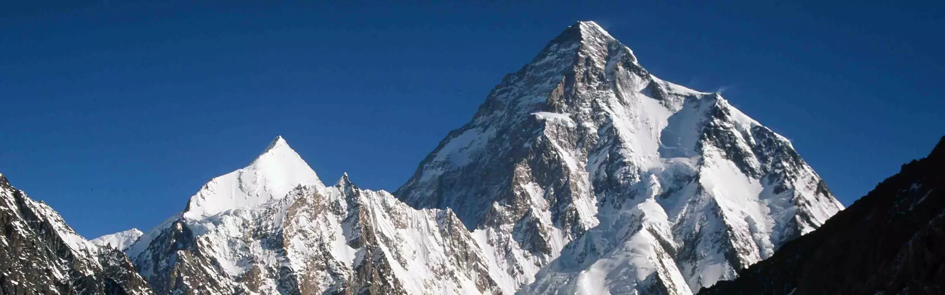Een bergbeklimmer in een rode jas en een gele broek staat op een besneeuwde bergtop en houdt een rood-wit-blauwe vlag met een embleem vast. De besneeuwde bergtoppen strekken zich uit tot in de verte o