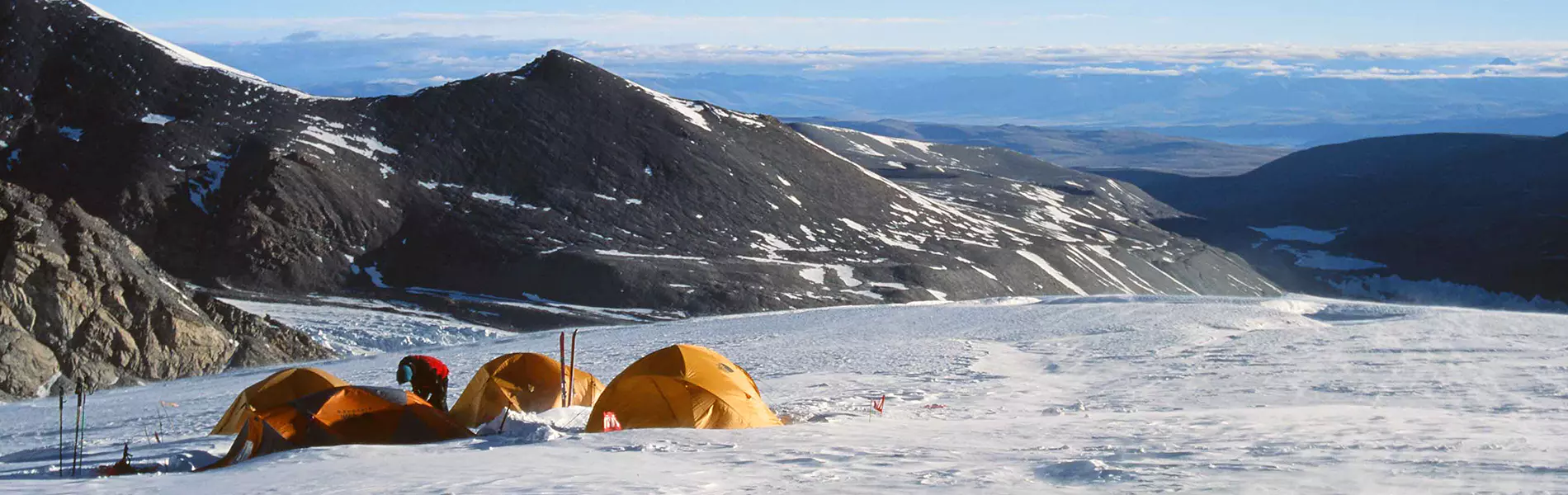Vier mensen in winterkleding wandelen door een besneeuwd berglandschap onder een strakblauwe hemel, omringd door met sneeuw bedekte bergtoppen en rotsachtig terrein.