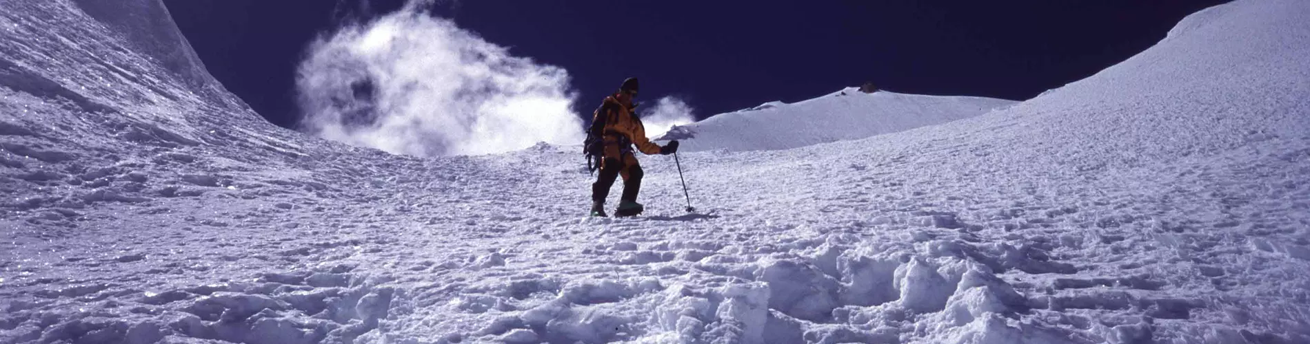 Een bergbeklimmer in een blauwe jas en een gele broek staat op de besneeuwde top van een berg en steekt triomfantelijk één arm in de lucht, met op de achtergrond besneeuwde bergtoppen en wolken onder 