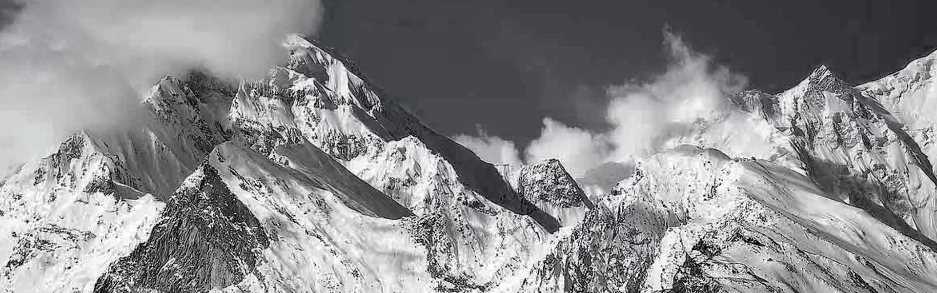 Met sneeuw bedekte bergtoppen onder een indrukwekkende hemel met grote wolken, vastgelegd in zwart-wit, waardoor de texturen en contrasten van het ruige landschap extra goed tot hun recht komen.