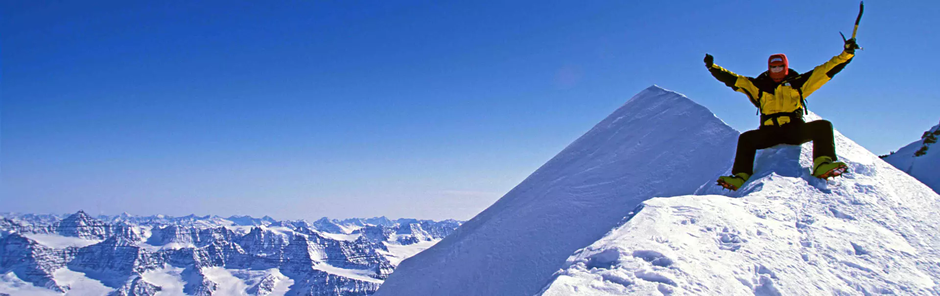 Een bergbeklimmer in een geel jack en een rode helm zit triomfantelijk op een besneeuwde bergtop, met opgeheven armen en een ijsbijl in de hand, tegen een helderblauwe lucht.