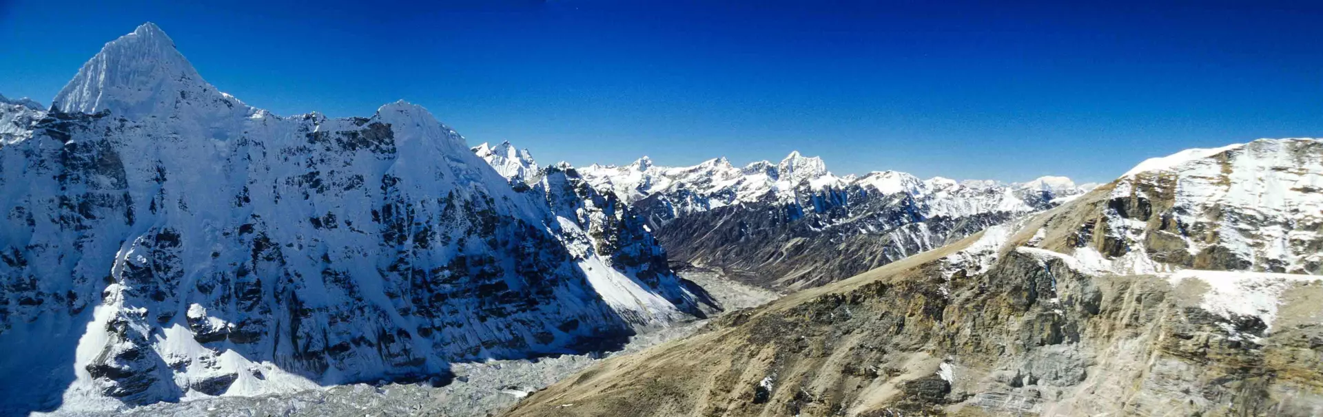 Een persoon in winterkleding staat op een besneeuwde bergkam, omringd door hoge, met sneeuw bedekte bergtoppen onder een helderblauwe hemel.