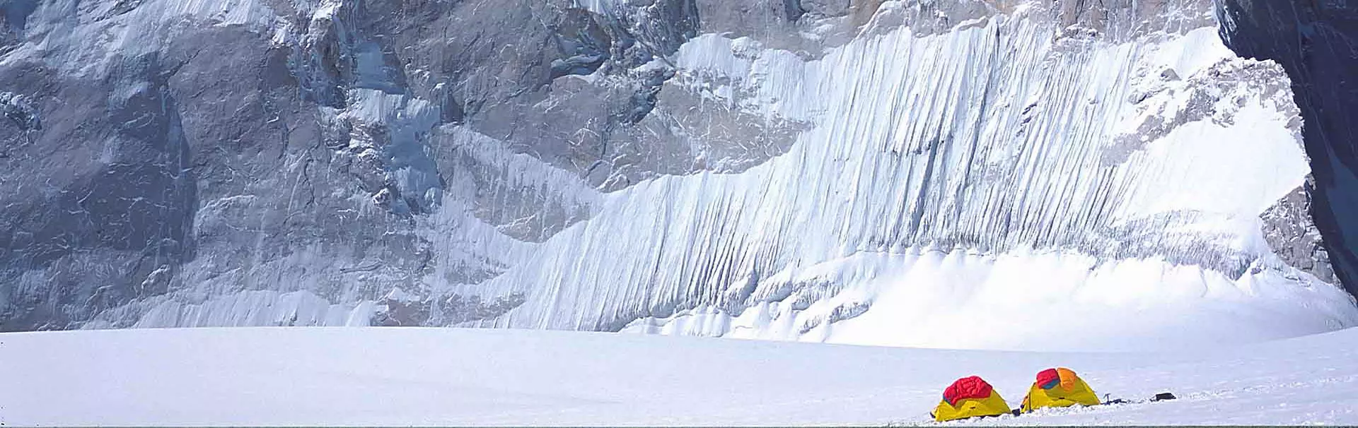 Iemand wandelt onder een helderblauwe hemel door een besneeuwd, ijzig landschap, op weg naar een torenhoge, ruige bergtop. Iets verderop rust een andere figuur uit bij een gletsjerspleet.