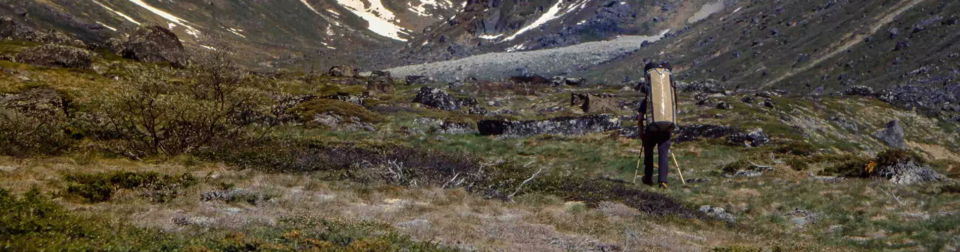 Twee klimmers met helmen op zitten in hangtenten die aan een steile rotswand zijn bevestigd, omringd door klimuitrusting. Op de achtergrond zijn besneeuwde bergtoppen te zien.