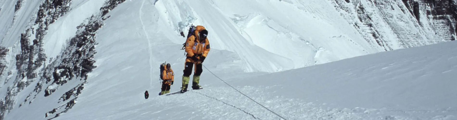 Een bergbeklimmer, gekleed in winterkleding en met een grote rugzak op de rug, beklimt een besneeuwde, steile berghelling, vastgemaakt aan een veiligheidstouw, met op de achtergrond ijzige bergtoppen 