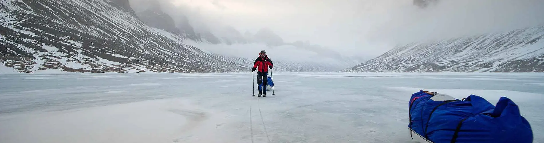 Twee wandelaars lopen door een rotsachtig, met sneeuw bedekt landschap, met op de achtergrond torenhoge kliffen en bergen, onder een helderblauwe hemel.