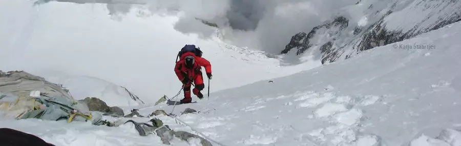 Drie bergbeklimmers in rode pakken en sneeuwuitrusting staan op een besneeuwde, rotsachtige helling vlakbij de top van een berg, waar ze onder een heldere hemel touwen en uitrusting gebruiken.