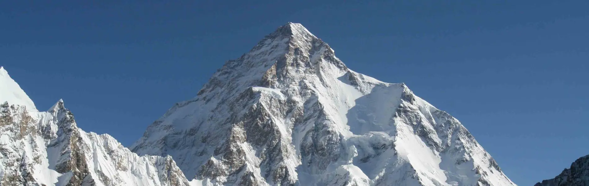 Vier bergbeklimmers in zware winteruitrusting staan op de besneeuwde top van een berg. Twee bergbeklimmers omhelzen elkaar uit vreugde, terwijl de anderen vlakbij staan, met fel zonlicht en een strakb