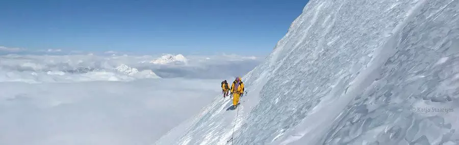 Een persoon met een bril en een gestreepte muts glimlacht terwijl hij geknield zit bij de ingang van een gele tent die op de sneeuw is opgezet; in de buurt liggen kampeerspullen, laarzen en een draagb