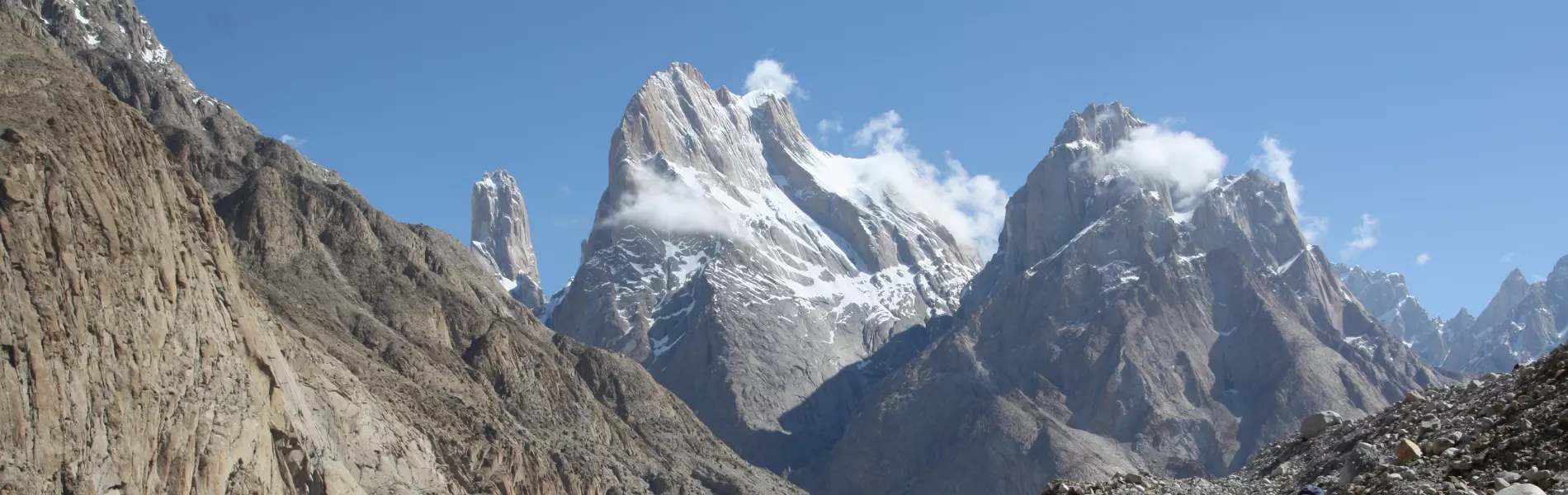 Een klimmer met een rugzak, waarvan alleen het silhouet te zien is, staat op een rotsrichel en kijkt uit over een indrukwekkend berglandschap met grillige, met sneeuw bedekte toppen onder een helderbl