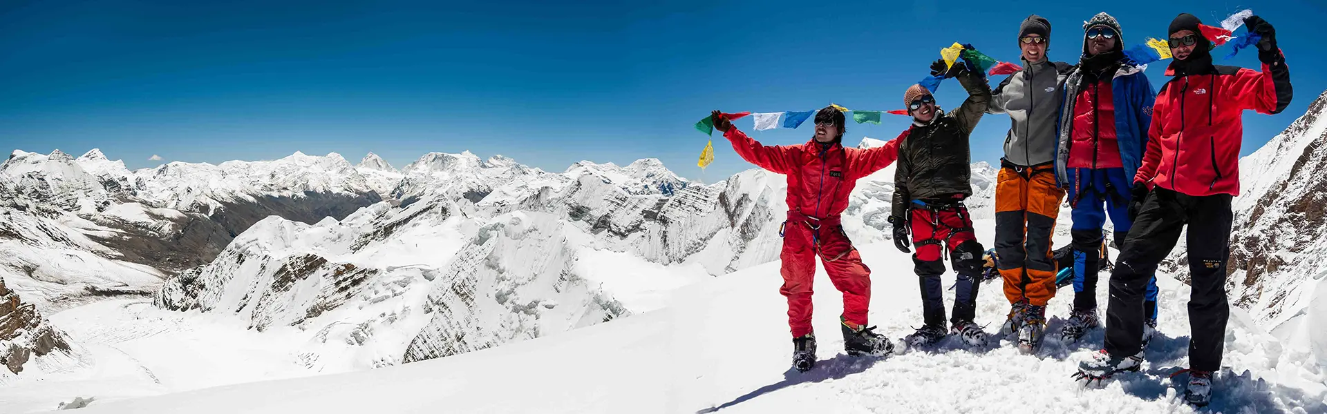 Een bergbeklimmer in rode winterkleding zit in de sneeuw, met naast zich klimuitrusting, touwen en een rugzak. Op de achtergrond zijn met sneeuw bedekte bergen en een bewolkte hemel te zien.