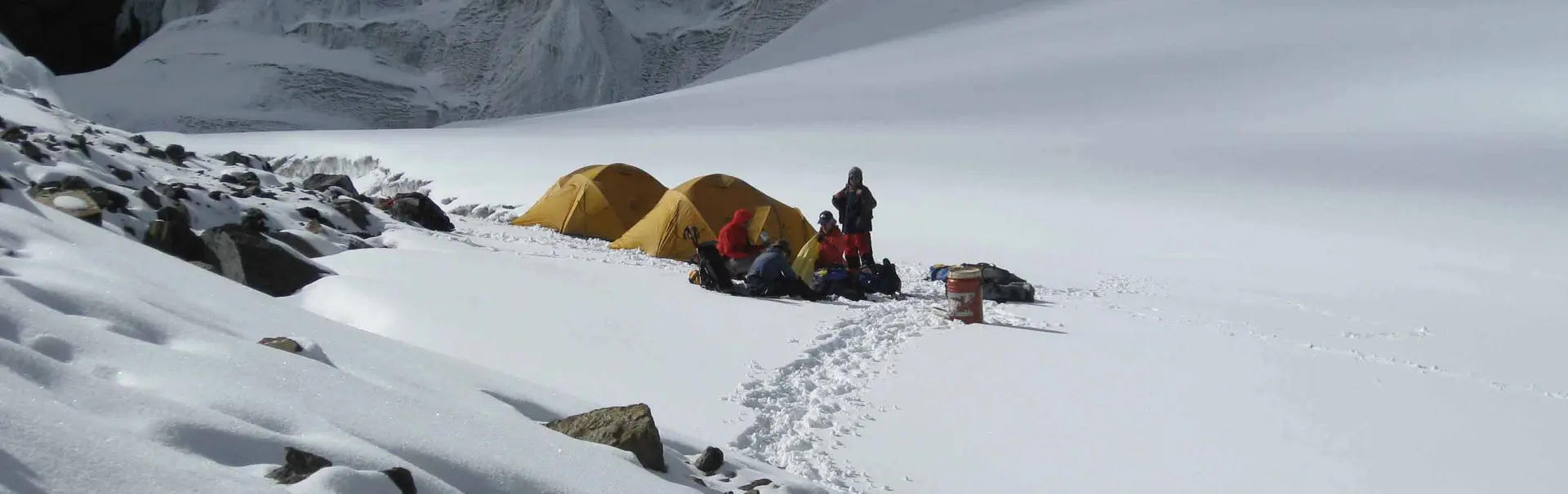 Een groep van zeven klimmers in bergbeklimmersuitrusting staat op een besneeuwde bergtop; ze glimlachen en poseren voor de camera, omringd door hun uitrusting en rugzakken, onder een heldere, gedeelte