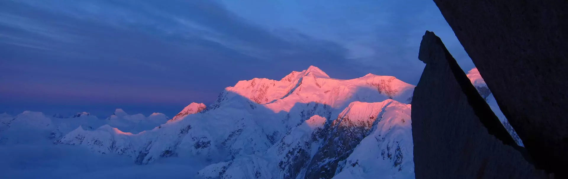 Vier glimlachende klimmers, gekleed in helmen, winterjassen en klimuitrusting, poseren dicht bij elkaar in de buitenlucht, met sneeuw en een blauwe lucht op de achtergrond. Eén van hen heeft een opger