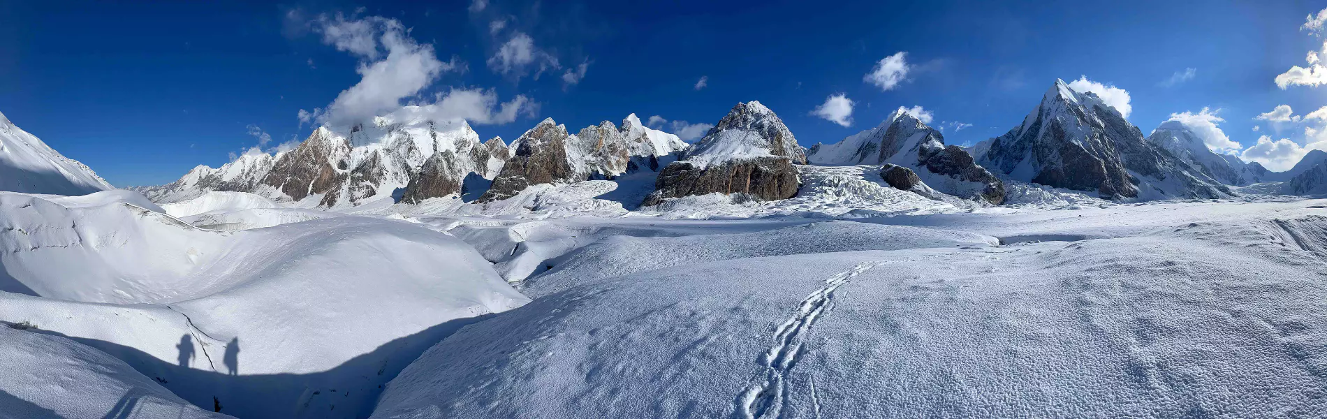 Twee bergbeklimmers met helmen en winterkleding glimlachen en houden een groene vlag vast op een besneeuwde bergtop, met op de achtergrond prachtige besneeuwde bergen en een blauwe lucht.
