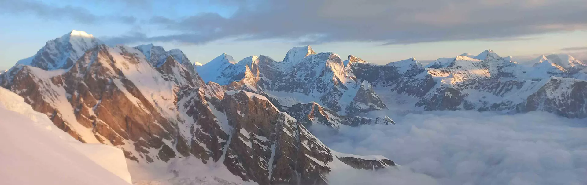 Een persoon met een zonnebril, een rode hoofdbedekking en een donkere jas glimlacht naar de camera terwijl hij in een besneeuwd berggebied staat, met wolken op de achtergrond.