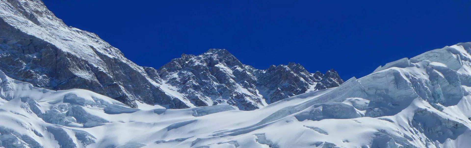 Twee mannen in feloranje winterjassen en winterkleding glimlachen voor een close-up-selfie op een besneeuwde berg, met een helderblauwe lucht en zonlicht dat weerkaatst in hun zonnebrillen.