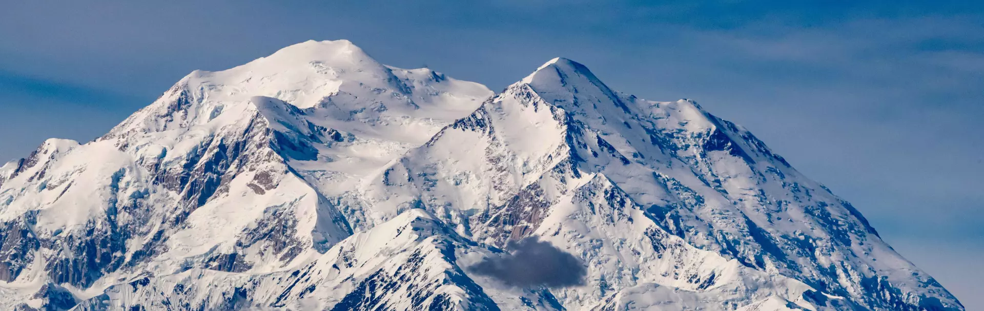 Een torenhoge, met sneeuw bedekte berg met ruige toppen en bergkammen steekt af tegen een blauwe lucht; op de lagere hellingen zijn sneeuwvlekken en rotsachtig terrein te zien.