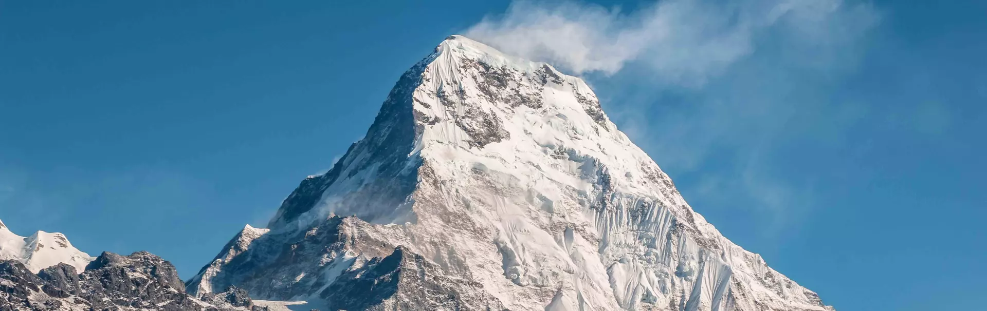 Een met sneeuw bedekte bergtop in fel zonlicht, met scherpe bergkammen en een helderblauwe lucht; een sneeuwwolk waait door de wind van de top af.