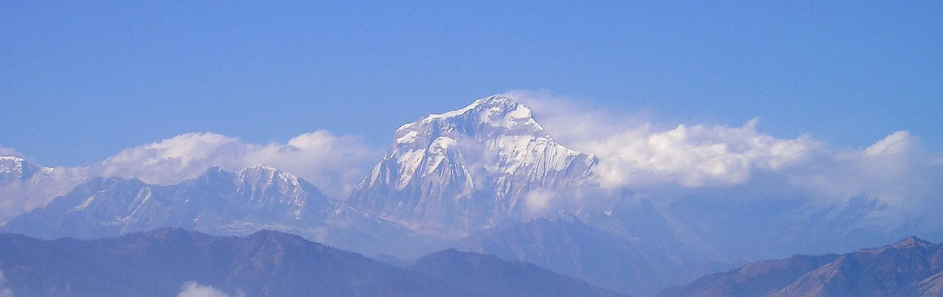 Een met sneeuw bedekte bergtop rijst boven de wolken uit, met een blauwe lucht op de achtergrond en mistige uitlopers op de voorgrond.