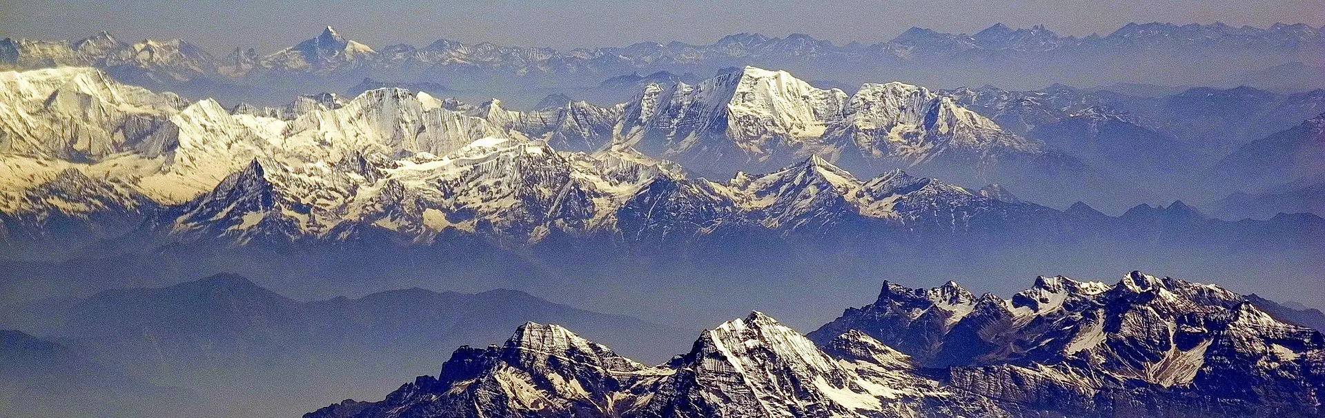 Een panoramisch uitzicht op de ruige, met sneeuw bedekte bergen van de Himalaya onder een helderblauwe hemel, met scherpe toppen en diepe valleien die zich tot in de verte uitstrekken.