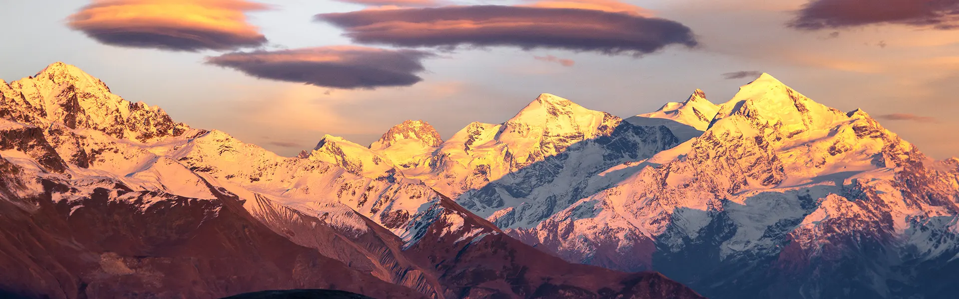 De met sneeuw bedekte bergtoppen gloeien in roze en oranje tinten in de zonsondergang, onder dramatische wolken aan een kleurrijke hemel. De donkere heuvels op de voorgrond vormen een contrast met de 