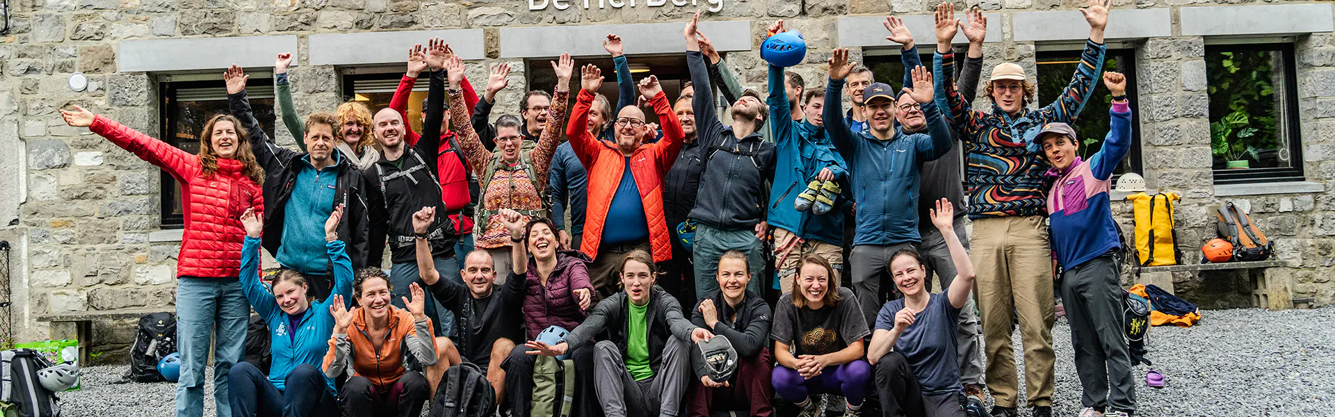 Een grote groep glimlachende mensen poseert voor een stenen gebouw, terwijl ze hun armen in de lucht steken en juichen. Sommigen dragen outdoor-kleding, wat doet vermoeden dat het om een wandel- of kl
