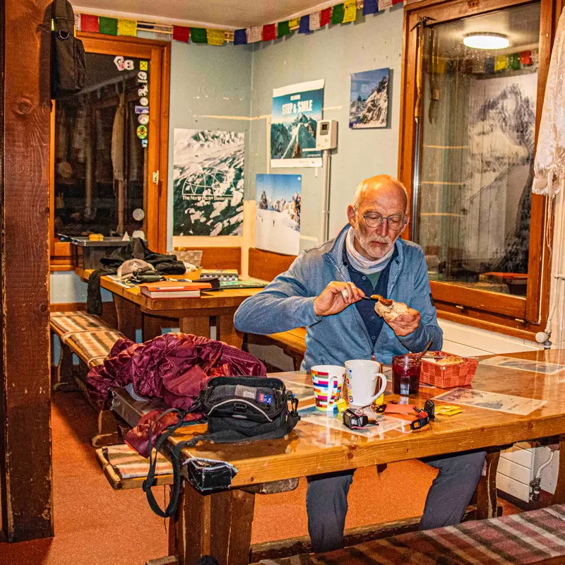 Een oudere man zit aan een houten tafel in een gezellige kamer en smeert jam op brood. Aan de muren hangen foto’s van bergen en gebedsvlaggen. Op de tafel staan mokken, een cameratas en ontbijtspullen.