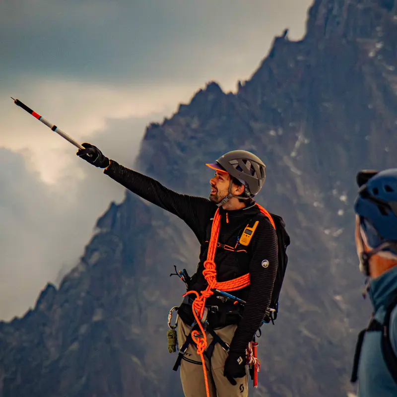 Een bergbeklimmer met een helm en een klimgordel richt een stok naar een rotsachtige bergtop, terwijl op de voorgrond dramatische wolken en een andere bergbeklimmer te zien zijn.