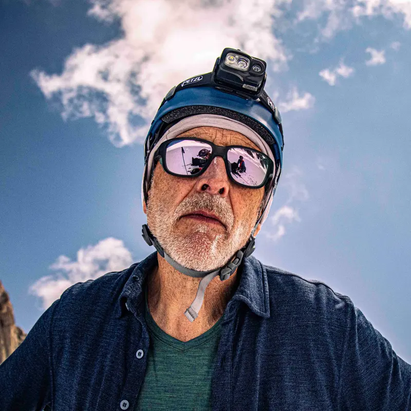 Een oudere man met een helm waarop een camera is bevestigd en een reflecterende zonnebril staat buiten onder een blauwe lucht met wolken en kijkt in de camera.