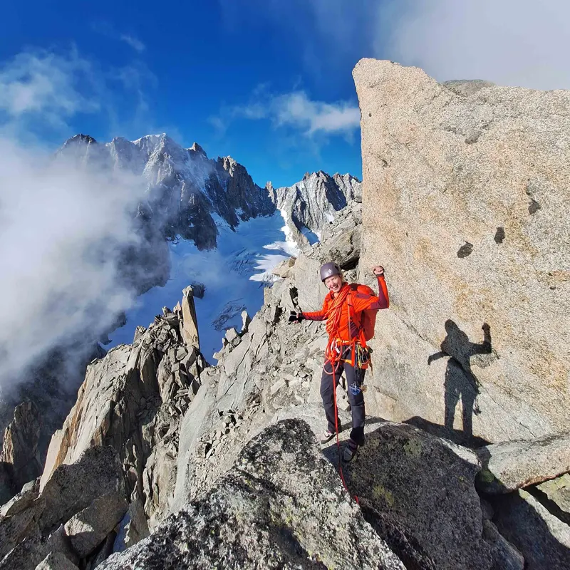 Een bergbeklimmer in een oranje jas staat op een rotsachtige bergkam, omringd door ruige bergtoppen en sneeuwvlekken, met op de achtergrond een blauwe lucht en wolken. De bergbeklimmer glimlacht en poseert met opgeheven armen.