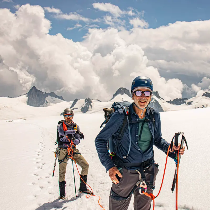 Twee bergbeklimmers met helmen en zonnebrillen, aan elkaar vastgebonden met een touw, staan in een besneeuwd berglandschap met dramatische wolken en grillige bergtoppen op de achtergrond. Ze zien er blij uit en lijken goed voorbereid op de omstandigheden in de bergen.