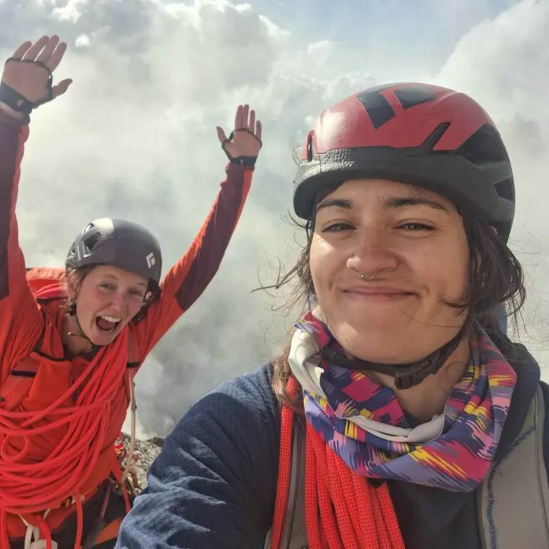 Twee klimmers met helmen en outdooruitrusting glimlachen naar de camera. De ene staat vooraan, terwijl de andere op de achtergrond opgewonden zijn armen in de lucht steekt. Ze worden omringd door wolken, wat doet vermoeden dat ze zich op grote hoogte bevinden.
