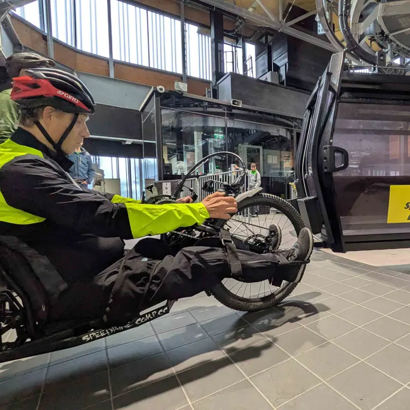 Een persoon in een ligfiets met een helm en fietsuitrusting wacht bij een open gondeldeur in een modern gebouw.