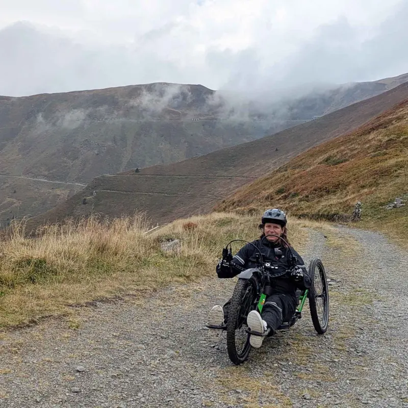 Een persoon met een helm op rijdt op een handfiets over een bergpad met grind, omringd door met gras begroeide heuvels en mistige bergen op de achtergrond.