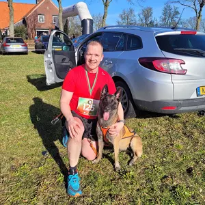 Een man in een rood shirt met een startnummer knielt naast een blije hond in een harnas; beiden glimlachen, voor een zilverkleurige auto en een grasveld op een zonnige dag.