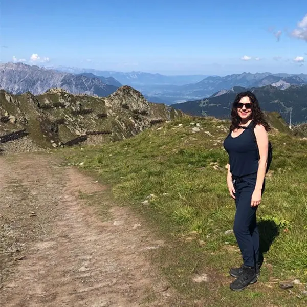 Een vrouw met lang donker haar en een zonnebril staat op een bergpad, omringd door met gras begroeide heuvels en rotsachtige bergen in de verte, onder een helderblauwe hemel.
