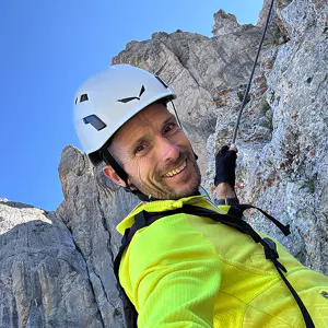 Een persoon met een witte helm en een geel jack glimlacht terwijl hij een rotsachtige berg beklimt, zich vasthoudend aan een touw, met een blauwe lucht op de achtergrond.