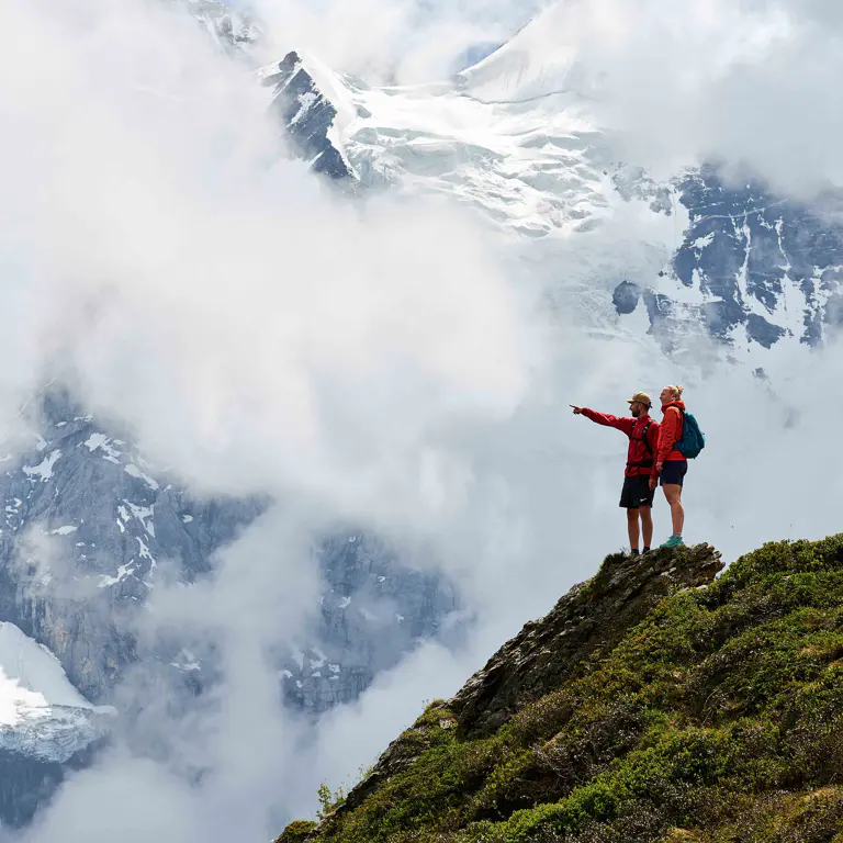 Twee bergwandelaars staan op een bergkam en wijzen naar een met sneeuw bedekte bergtop.