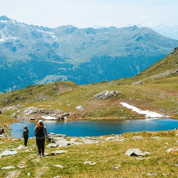 Twee mensen met rugzakken wandelen naar een klein bergmeer, omringd door met gras begroeide heuvels en rotsachtig terrein, met in de verte een reeks bergen tegen een blauwe lucht.