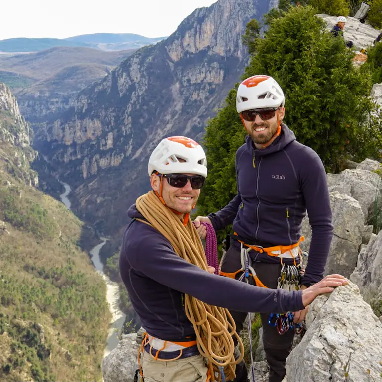 Twee bergbeklimmers met helmen en klimgordels poseren op de rand van een klif die uitkijkt over een rivierkloof.
